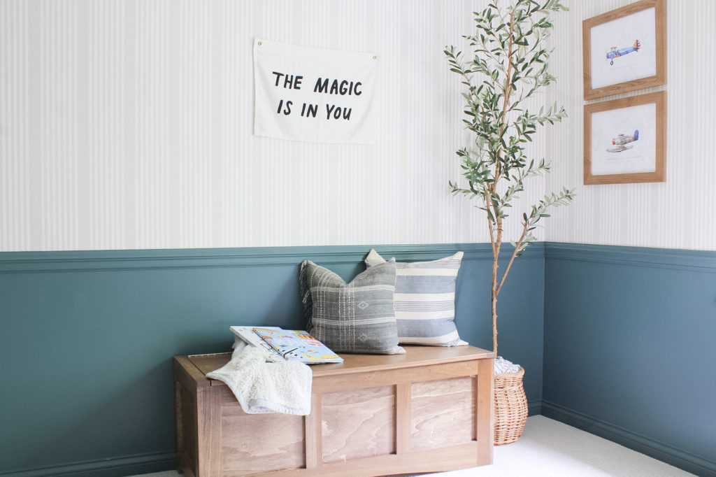 An airplane themed boy's bedroom design. Walls are painted sherwin william's riverway. There are grey Jenny Lind bed's. A tan stripe wallpaper is on the walls. Vintage airplane artwork hangs on the wall. 