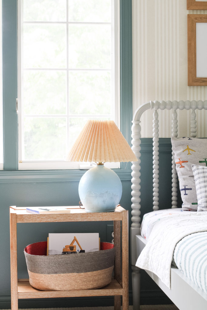 A Crate & Barrel Gray Jenny Lind bed is next to a Serena & Lilly wood side table with a blue lamp on top. The walls are painted sherwin william's riverway on the lower half and above the chair rail is a linen stripe wallpaper. 