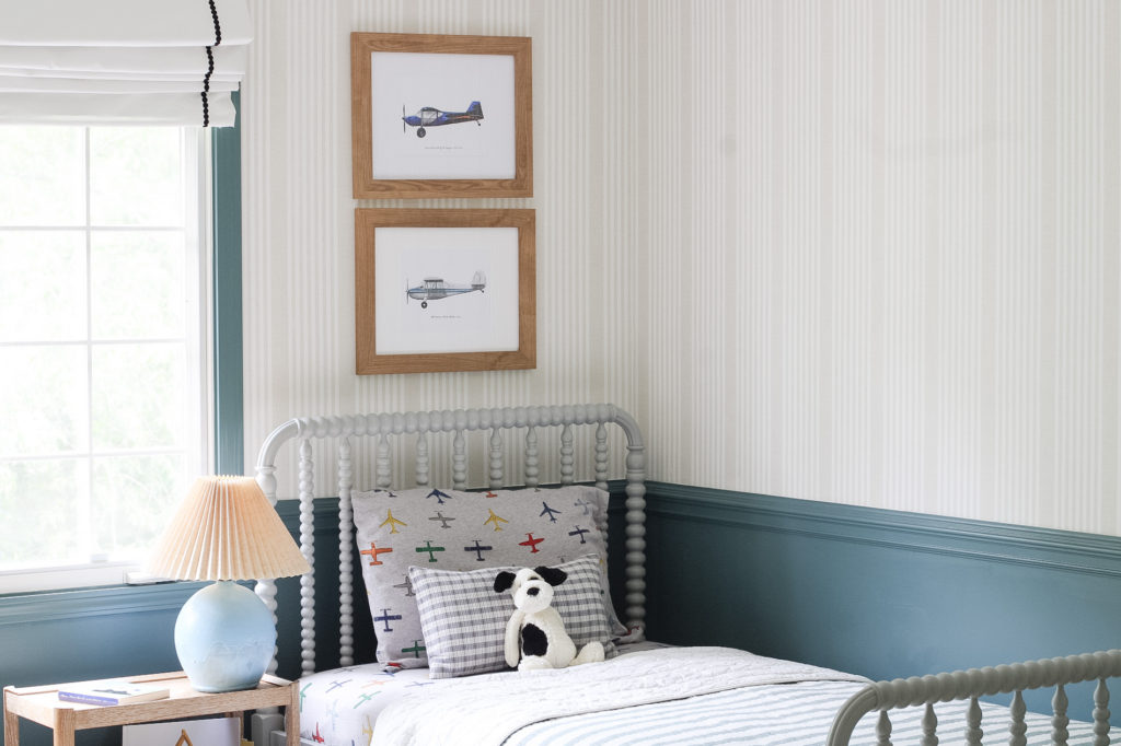 An airplane themed boy's bedroom design. Walls are painted sherwin william's riverway. There are grey Jenny Lind bed's. A tan stripe wallpaper is on the walls.  Airplane illustrated artwork is on the walls. 