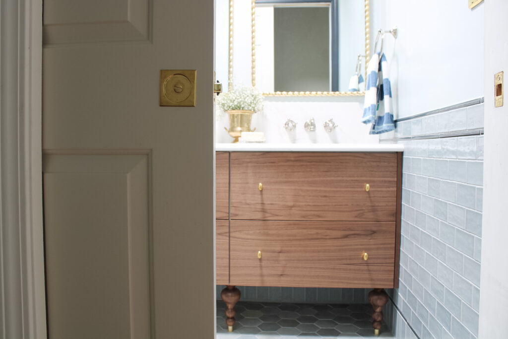 Pocket door tubular hardware in unlacquered brass opening a door into a blue tile bathroom with a walnut vanity and brass rope framed mirrors