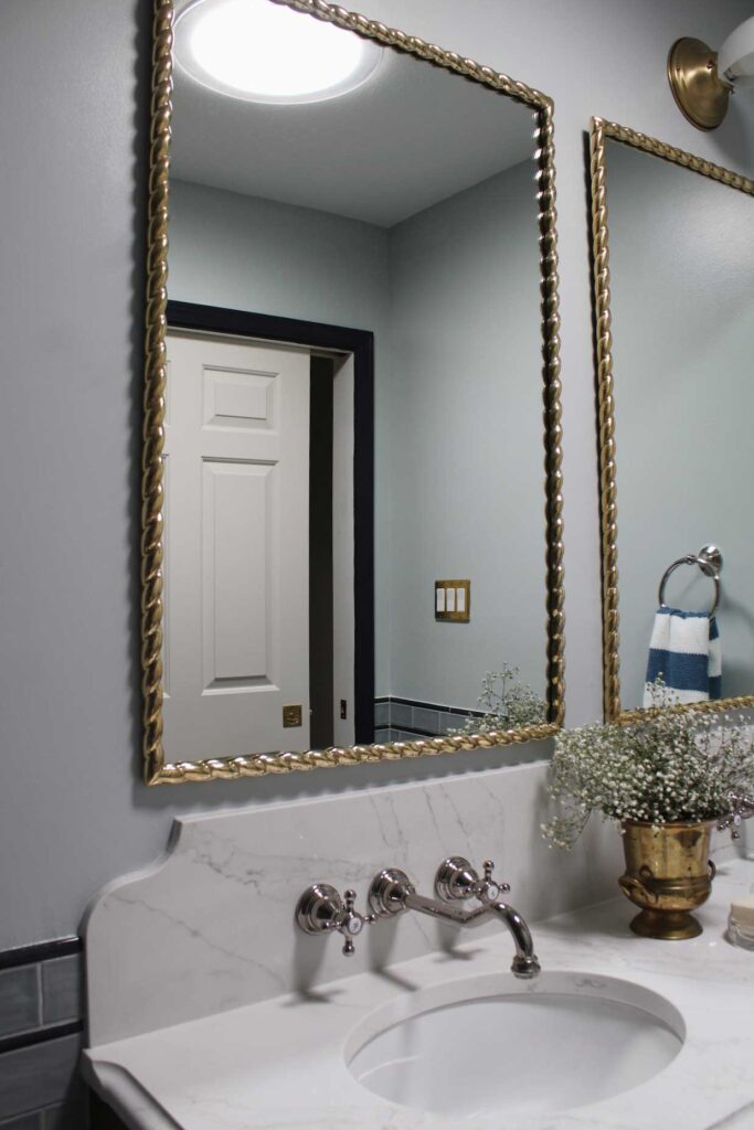 Brass mirrors hang over the walnut bathroom vanity. Wallmount faucets are in a vadara sterling light quartz backsplash. Blue subway wall tile is on the wall.