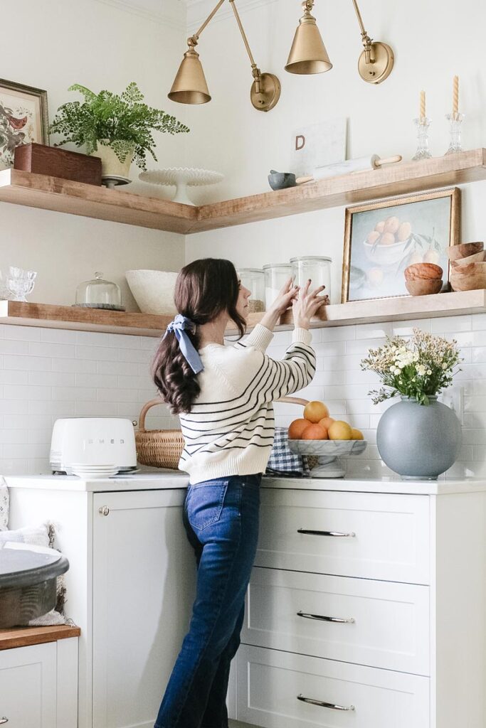 an interior designer is styling open wood kitchen shelves above white cabinets in a kitchen. 